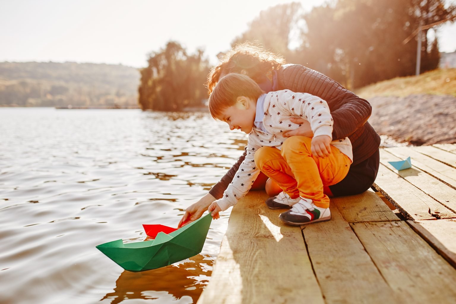Eine Frau mit Kind sitzt auf einem Steg und spielt mit einem Papierboot im Wasser